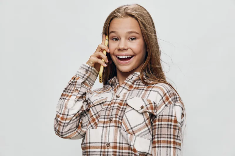 Girl with long hair smiling and holding smartphone near ear for phone call communication. Portrait, casual clothing and positive expression on gray background.