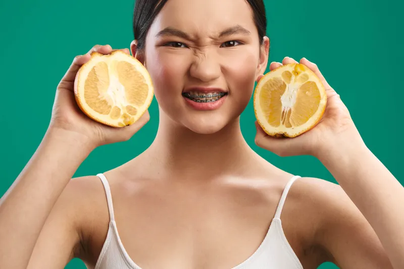 Asian woman holds halved lemons near her face, smiling playfully against a vivid green studio backdrop, conveying freshness, energy, and a bright, natural vibe.