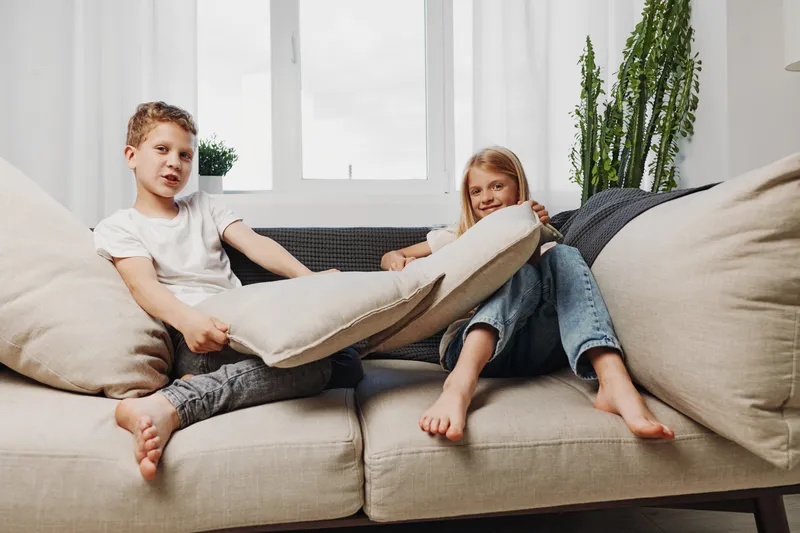 Two barefoot children having fun on beige sofa, playfully fighting with large pillows in bright living room with plants and natural light from window behind.