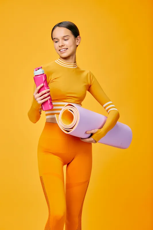 Fitness woman in bright orange activewear holds a pink water bottle and a rolled yoga mat against a vibrant orange background, ready for workout and sport sessions.