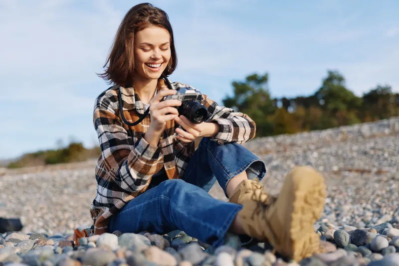 Photographer in a casual outfit captures a smiling moment on a rocky beach, outdoor scene with camera in hand, showcasing lifestyle, nature, and relaxed exploration against a sunny seashore.