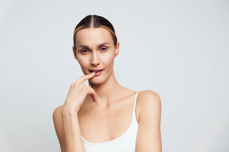 Young woman with clear skin and slicked back hair poses with finger near lips looking at camera wearing white tank top against plain light background.