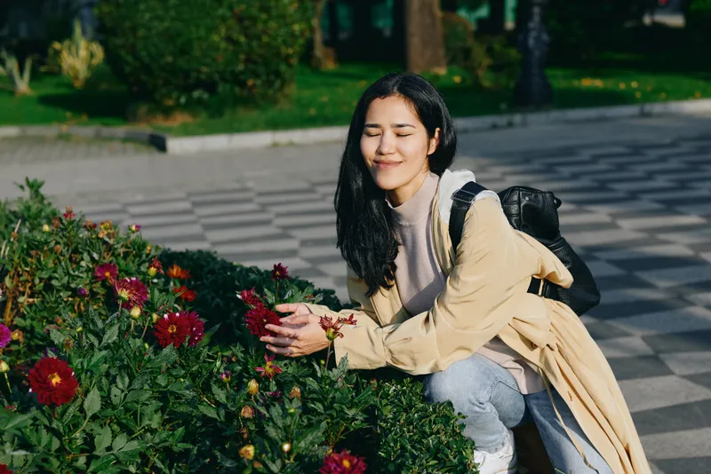 A woman kneeling beside vibrant flowers in a garden, smiling softly as she admires blossoms, wearing a light coat and backpack, enjoying a sunny outdoor moment.