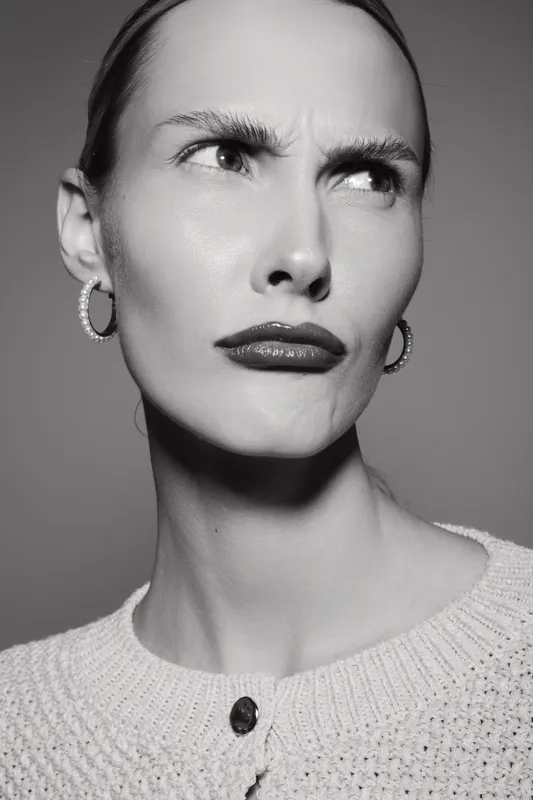 Close-up portrait of a serious slender white woman with pale skin and slicked-back hair wearing hoop earrings and a sweater. Concept of emotion, expression, and fashion.