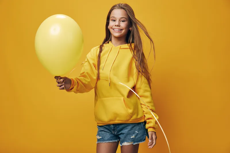 Young girl in yellow hoodie holding yellow balloon smiling standing against yellow background studio portrait with casual denim shorts and long hair.