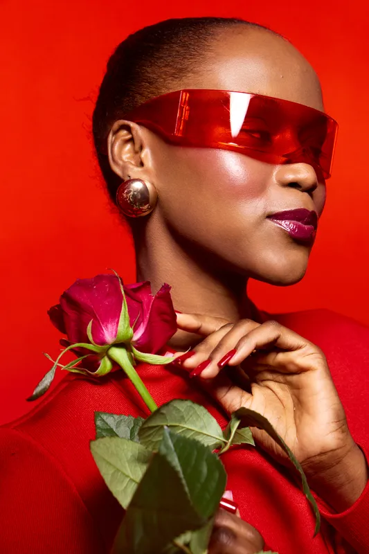 Stylish black woman with dark skin wearing red futuristic sunglasses and earrings holds red rose against bright red background. Concept of fashion, elegance, and beauty with floral elements.