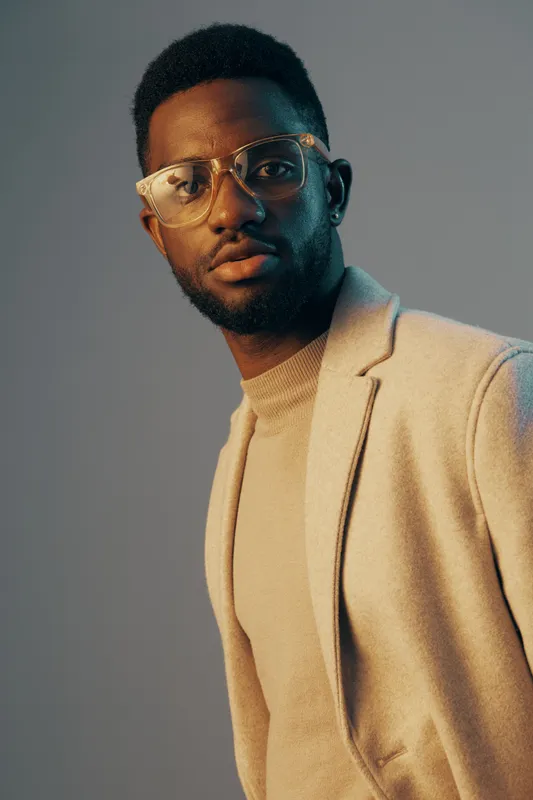 Stylish African man with glasses in a soft beige blazer, exuding confidence and sophistication against a pastel gradient background, ideal for fashion and portrait concepts