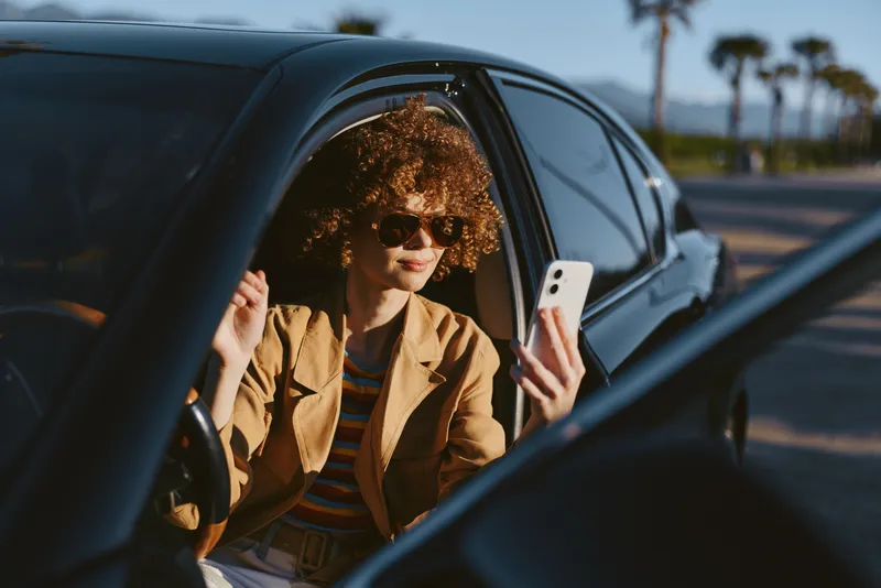 Casual woman in a car, smiling at a smartphone, sunlit moment with retro film color, relaxed outdoors vibe and natural lifestyle details