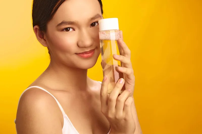 Asian woman gracefully presents a skincare product in a bright studio, showcasing smooth skin and a confident smile while holding a bottle against a yellow backdrop, conveying beauty, wellness, and