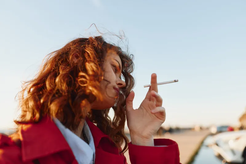 Young woman with curly hair wearing a red coat smokes a cigarette outdoors during daytime with a clear sky background.