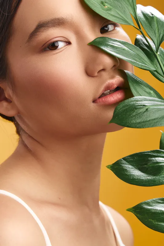 asian woman is presented in a closeup portrait, with glossy plant leaves partially shading her face against a warm yellow background, creating a serene, natural mood and soft, flattering lighting.