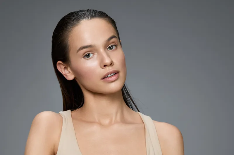 Natural beauty portrait of a young woman with minimal makeup, featuring healthy skin and wet hair against a grey background, emphasizing simplicity and elegance