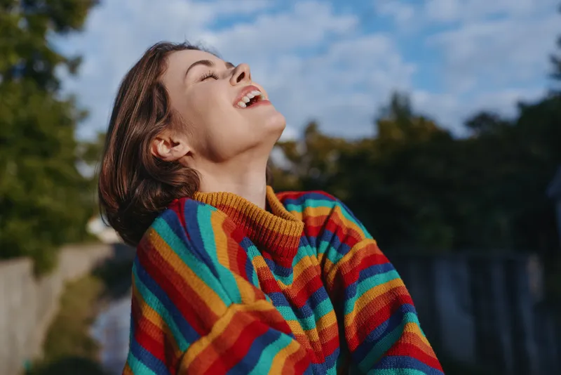 Woman portrait with laughter in a colorful sweater, joyful outdoor scene showing happiness and cozy autumn vibe, smiling young adult tilting head back in a candid knitted jumper moment.