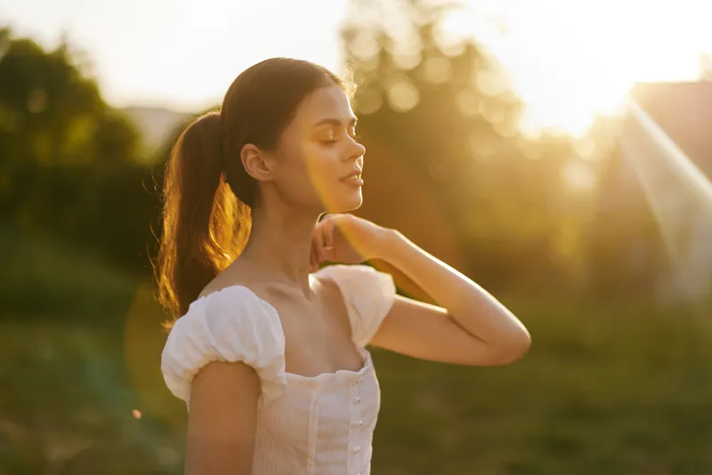 Young woman in a white dress enjoying a sunset in a green field, radiating peace and tranquility with a glowing sun background