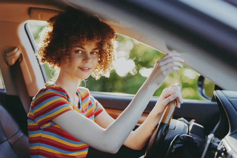 Young woman drives a car, smiling at the camera, wearing a striped tee, vintage film colors, natural sunlight, casual road trip mood, focused on the road and feeling free, and spontaneity.