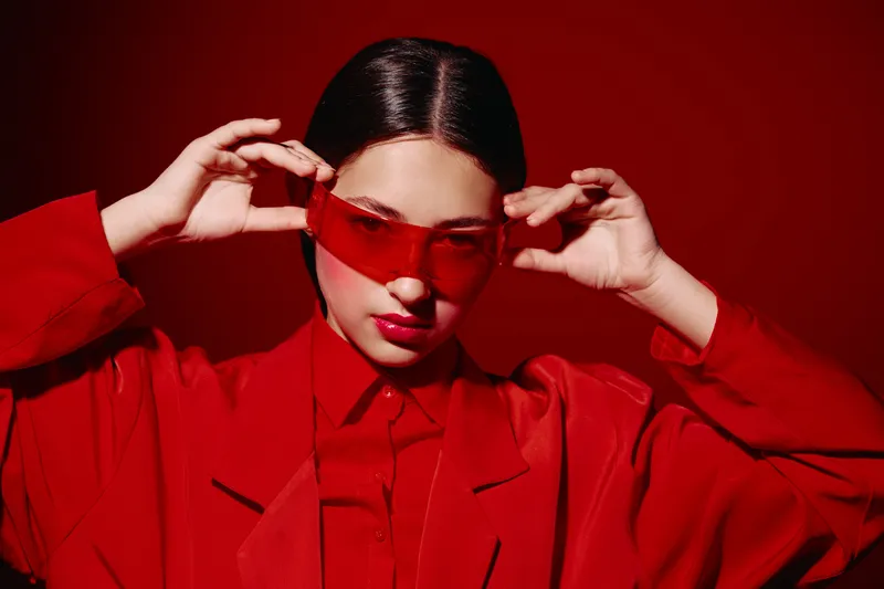 Stylish young woman wearing oversized red glasses and red high fashion clothing adjusts her sunglasses against a stark red background. Concept of beauty, creativity and fashion.