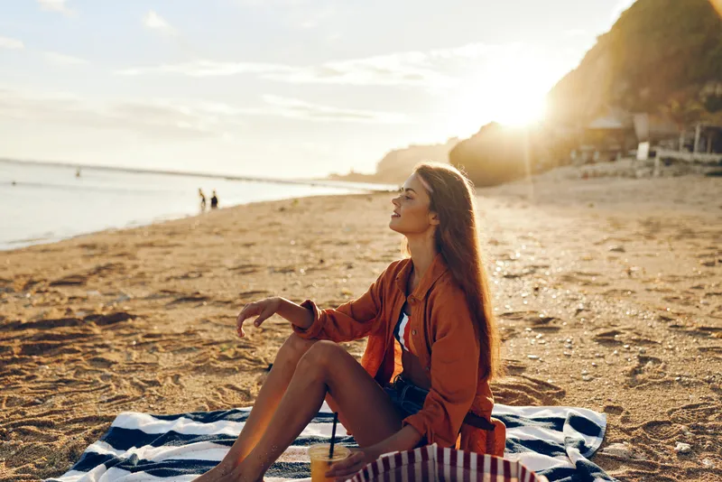 A young woman sits on a striped towel at a sunlit beach, wearing a brown jacket, gazing toward the horizon as warm light paints the water and sand.