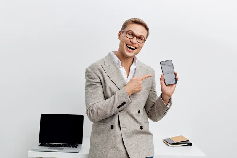 excited young caucasian man wearing beige casual blazer pointing smartphone screen with finger stand near laptop office desk isolated on white background remote work digital nomad concept