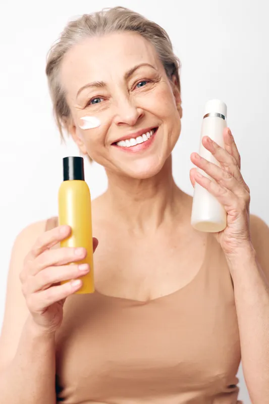 Smiling mature woman with short hair holds two cosmetic bottles and applies white cream on cheek. Bright studio background. Concept of skincare, beauty routine, self-care, and healthy aging.