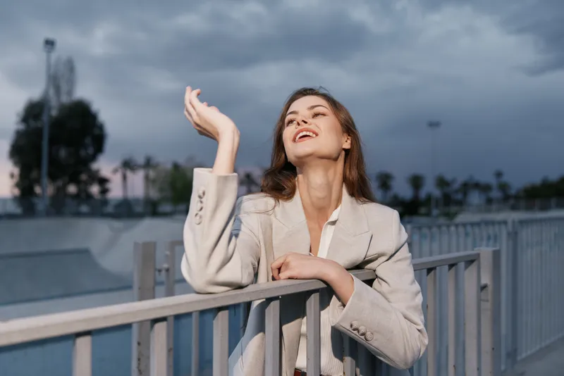Woman in light blazer expressing joy and confidence outdoors with cloudy sky background, natural lighting, casual style, and carefree emotions