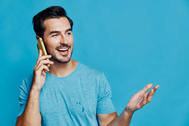 Smiling man talking on mobile phone with cheerful expression, wearing casual blue t-shirt, set against a solid bright blue background. Communication lifestyle concept
