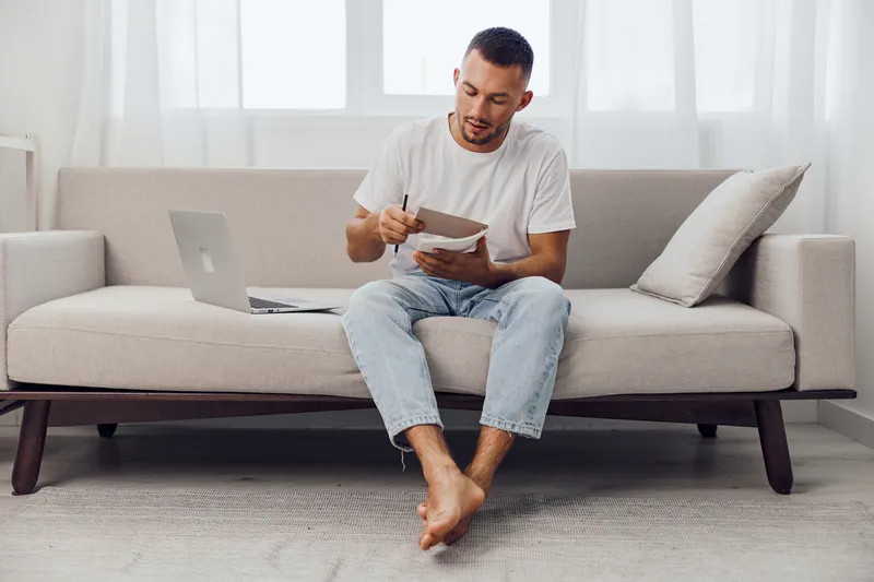 Thoughtful man brainstorming ideas at home, sitting on a cozy couch with laptop and notebook, creating content in bright and modern interior space.