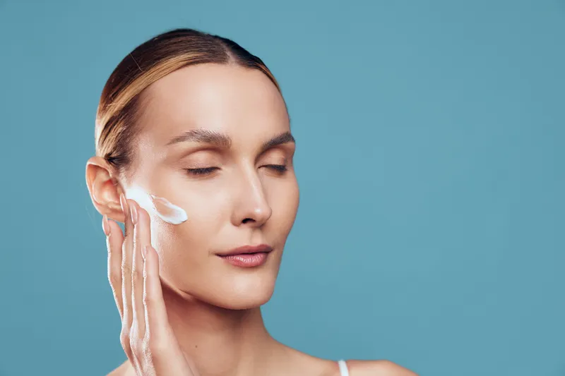 A young adult blonde woman with a calm expression applies moisturizer to her face in a studio with a blue background, showcasing a serene skincare moment and self care ritual.