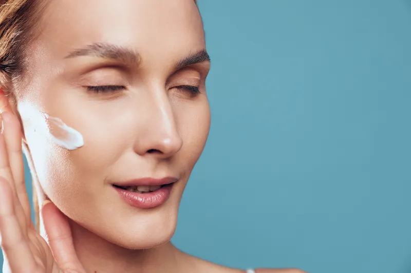 A young woman with closed eyes applies white cream on her cheek against a blue background. Concept of skincare, beauty routine, and facial care for healthy skin.