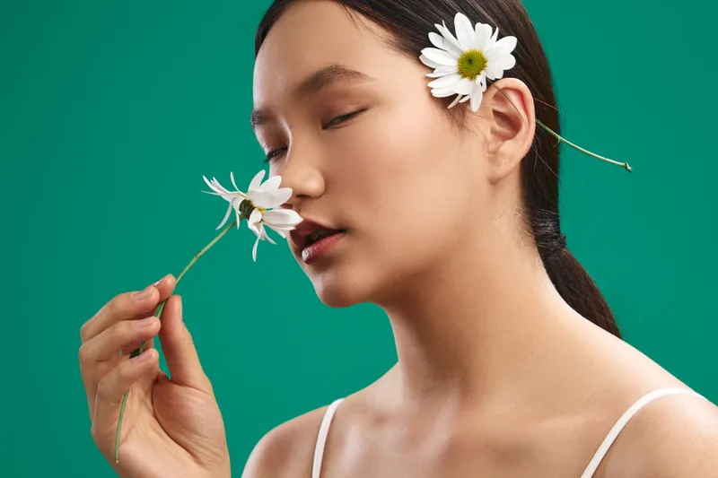 asian woman with flowers in her hair smiles softly, presenting a serene natural portrait in a minimal studio setup with a vivid green backdrop, radiating calm, grace, and understated elegance.