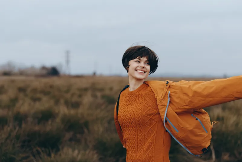 Smiling woman lifestyle outdoors in a grassy field wearing orange sweater and jacket, wind lifting her hair as she faces the camera, calm candid moment in nature during autumn sunset light