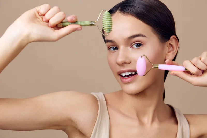 Young woman smiling while using facial rollers for skincare, showcasing natural beauty and wellness, in a soft beige background