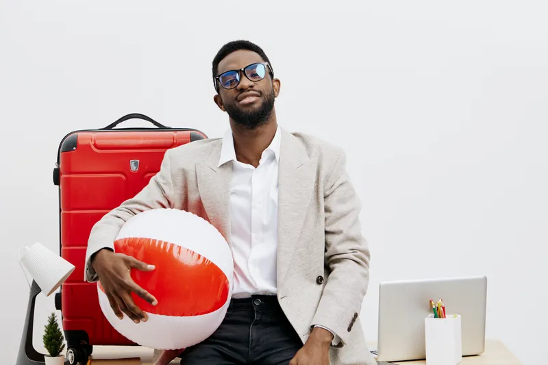 Young stylish man with sunglasses dressed in beige blazer and white shirt holding a colorful beach ball in front of travel luggage and laptop on a white background. People lifestyle concept