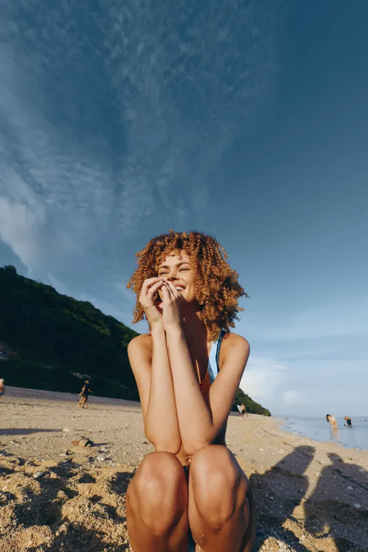A young woman with curly hair sits on a sunny beach in a blue swimsuit, smiling softly with her hands near her face as waves shimmer behind her.