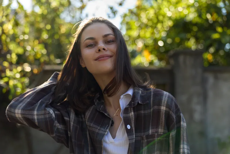 A smiling woman in a plaid shirt stands outdoors, capturing a relaxed lifestyle moment in a sunny park, showcasing casual confidence and natural charm conveying warmth