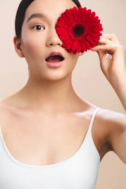 Asian woman holds a bright red flower beside her eye, soft smile, minimalist studio backdrop, creating a graceful and confident contemporary portrait with a vibrant focal point.