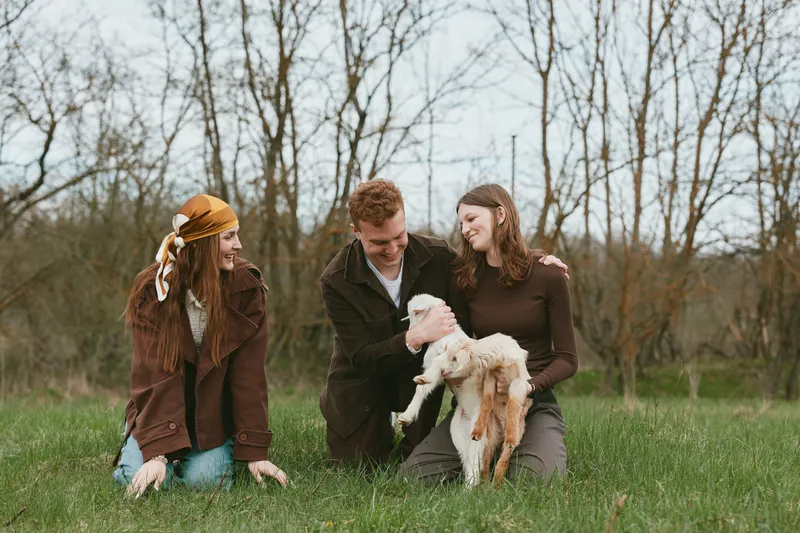 Three friends share a sunny afternoon in a grassy field, gently cradling a small goat kid. Warm, candid moment highlighting countryside lifestyle, friendship, and animal bonding.
