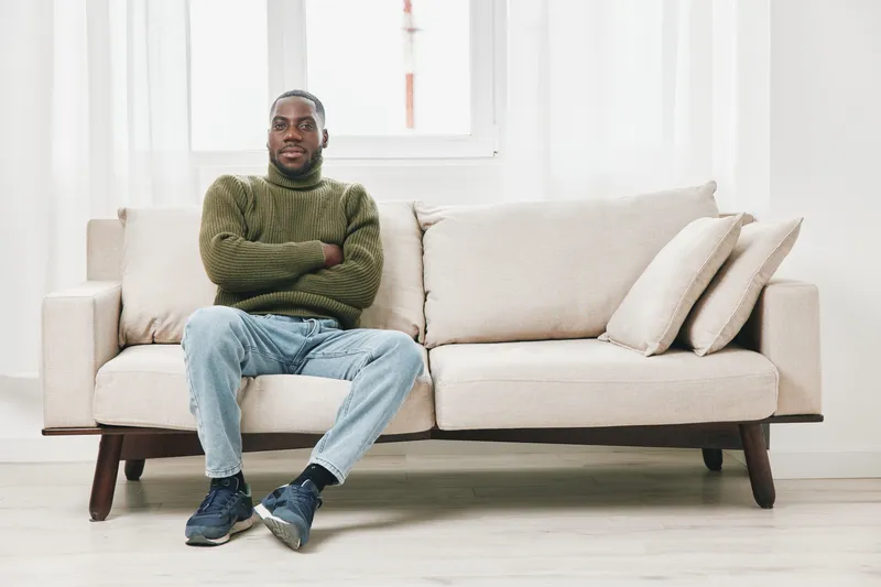 A relaxed African American man in a cozy green sweater sits comfortably on a beige sofa, expressing calmness and confidence in a bright, minimalistic room