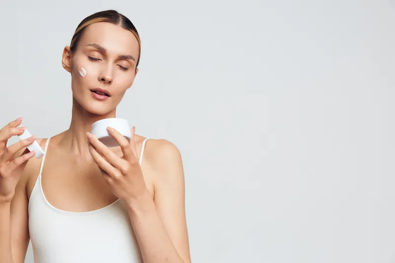 Young woman with closed eyes applying white face cream on cheek, holding jar and spatula. Minimalist background, concept of skincare, beauty routine, and self-care with soft lighting.