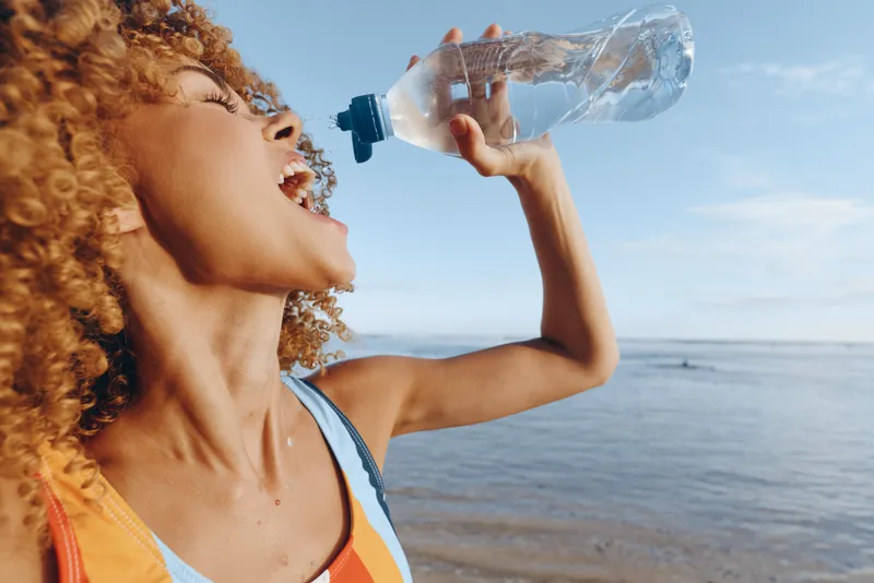 Young african american woman with curly hair drinks water from a plastic bottle while standing near the sea on a sunny day, enjoying hydration and outdoor activity.