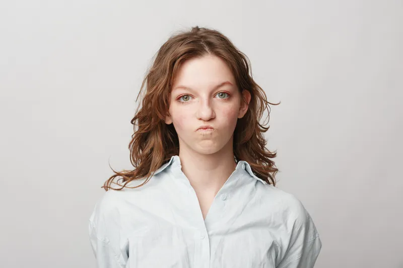 Young woman with curly hair makes a funny face with pursed lips and raised eyebrows. Casual style on a plain light background. Concept of humor, expression, and personality.