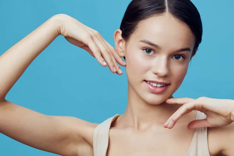 A young model with fair skin and a natural expression poses gracefully against a soft blue background, showcasing elegance and beauty with a focused gaze and gentle hand movements