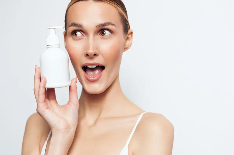 Young woman with clear skin holding a white pump bottle near her face, looking surprised. Concept of skincare, beauty, health, and cosmetic product. Bright studio lighting on a white background.