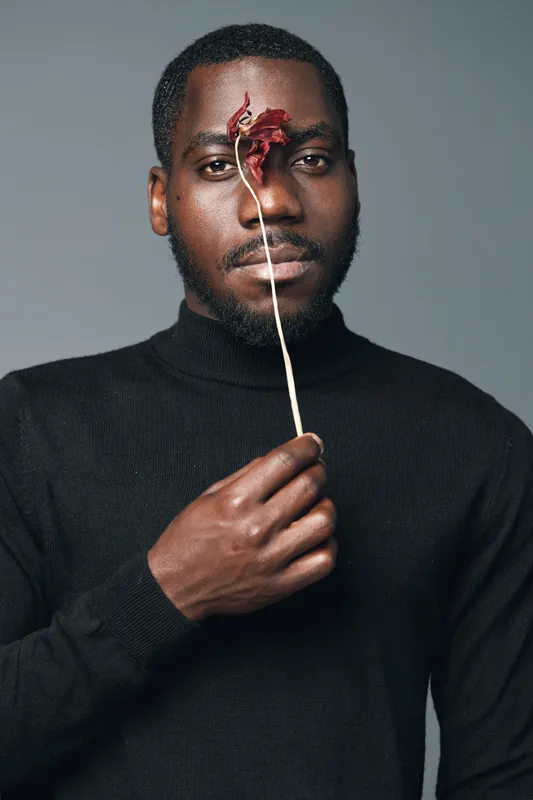 African man with dark skin wearing black sweater holding dried flower in front of face in creative conceptual visual style studio portrait. Artistic, minimalistic and modern expression.