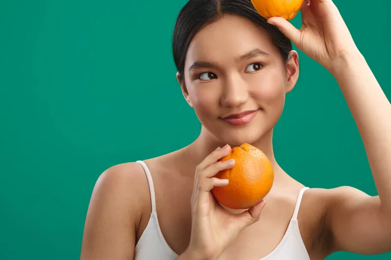 Asian woman is captured in a bright studio, holding oranges near her head and chin with a playful, curious smile, creating a fresh, vibrant scene that suggests health and creative energy.
