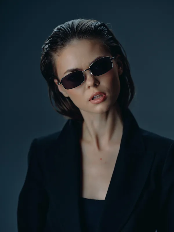 Stylish woman wearing sunglasses and black jacket with moody expression on dark background, fashion portrait concept.