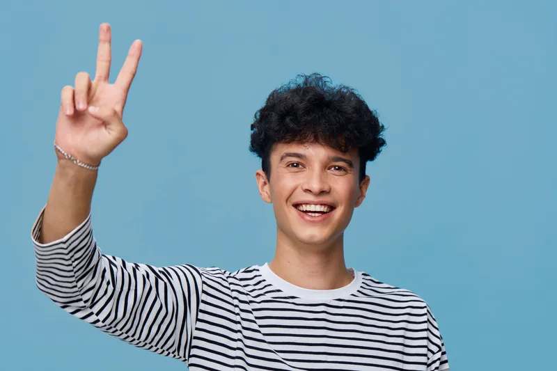 smiling young man in striped shirt showing peace sign with happy expression against blue background, casual style and positive mood
