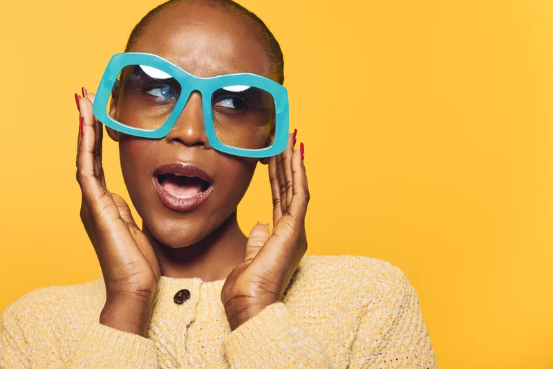 Portrait of a surprised African American woman with dark skin wearing large blue sunglasses and a beige sweater on yellow background. Concept of fashion, expression, and accessories.