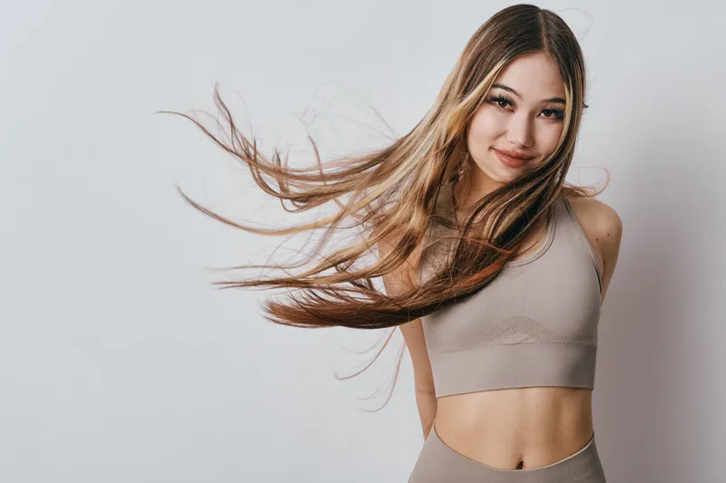 Young woman with long hair and sportswear posing with hair blowing, smiling confidently against a neutral background for fitness, wellness, and lifestyle concepts.