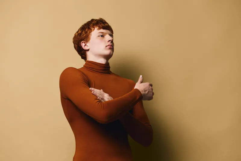 Young man with red hair wearing brown turtleneck, posing with crossed arms, looking confident against beige background in studio. Portrait of thoughtful male model with serious expression.