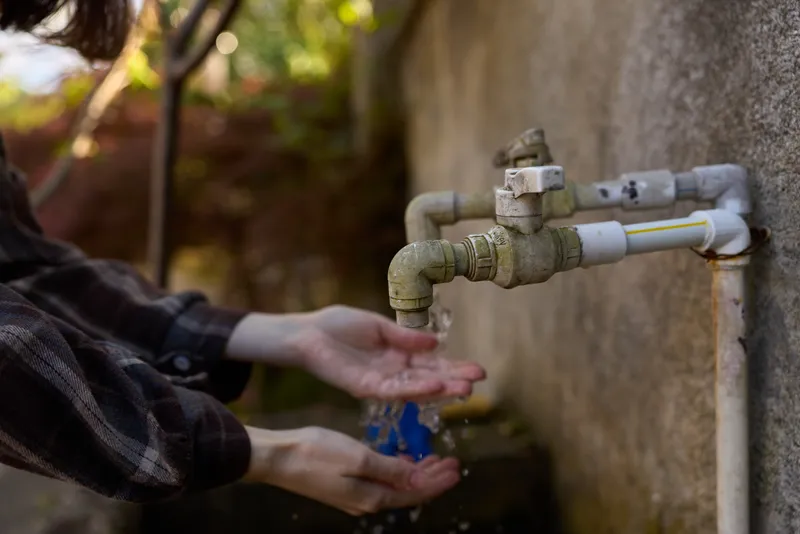 Young woman washes her hands under an outdoor wall faucet, wearing a plaid shirt, capturing a practical everyday moment of hygiene and calm